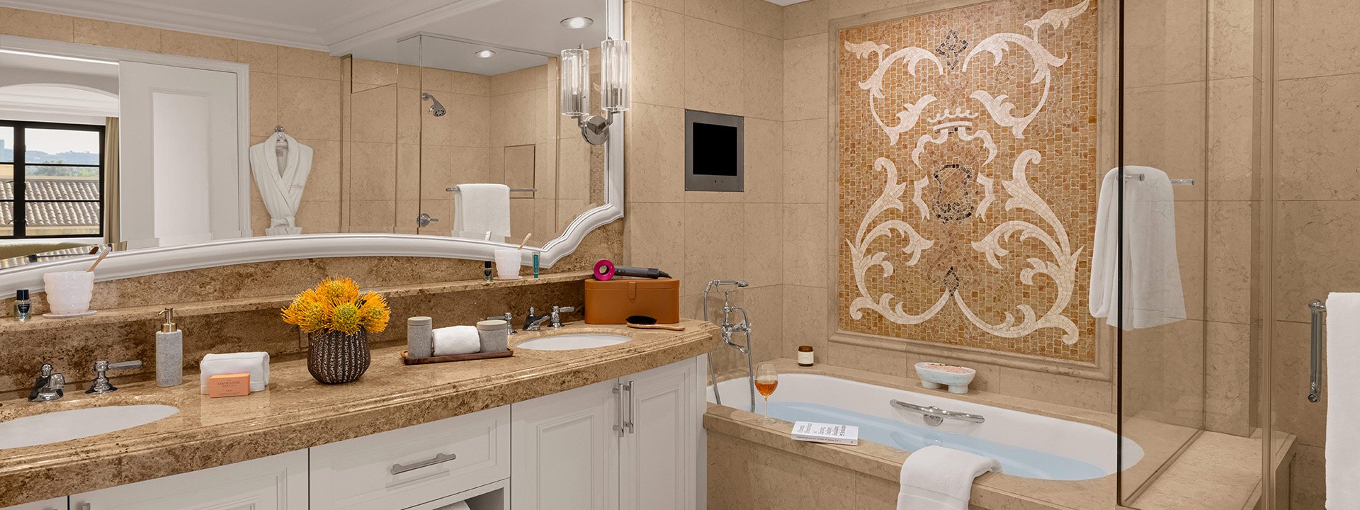The bathroom of a hotel room with two sinks and a large bathtub. There is a mosaic wall above the bath tub.
