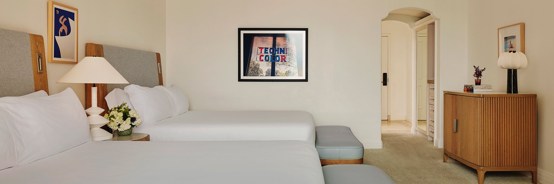Two queen beds with white sheets, a small wooden credenza, and a nightstand with white flowers and a white lamp.