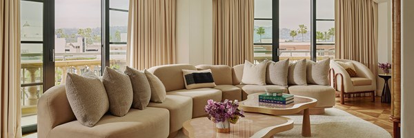Large L-shaped beige sofa with beige cushions in living area of suite. There are potted purple flowers and books on a table.