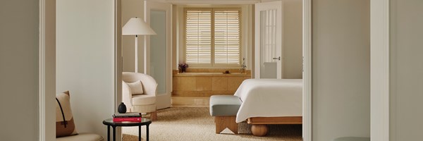 A bed with white sheets, a glass table with books, and a sitting chair. Doors are open to a bathroom and bathtub.