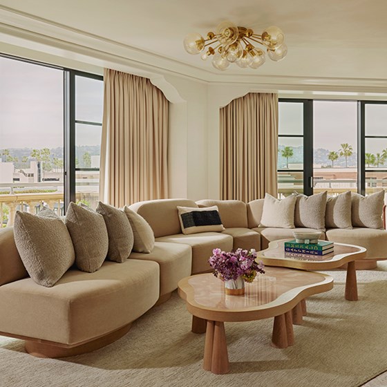 Large L-shaped beige sofa with beige cushions in living area of suite. There are potted purple flowers and books on a table.