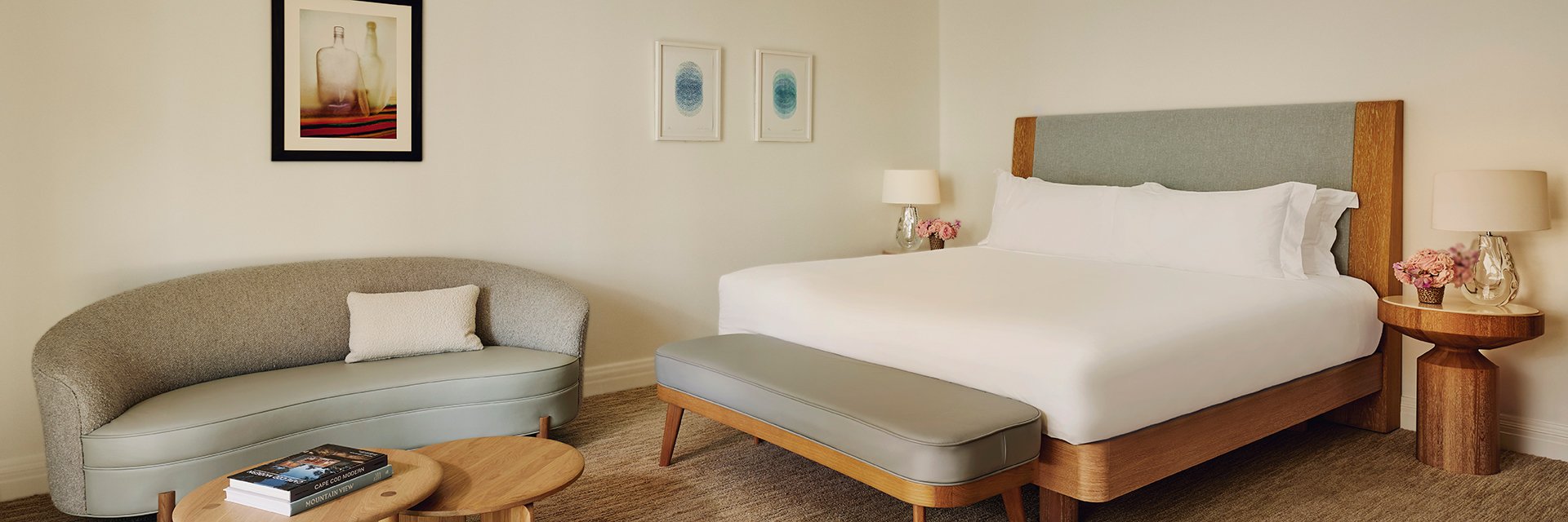 A neat king's sized bed with white sheets, two wooden nightstands with pink flowers and lamps, a coffee table, and a sofa.