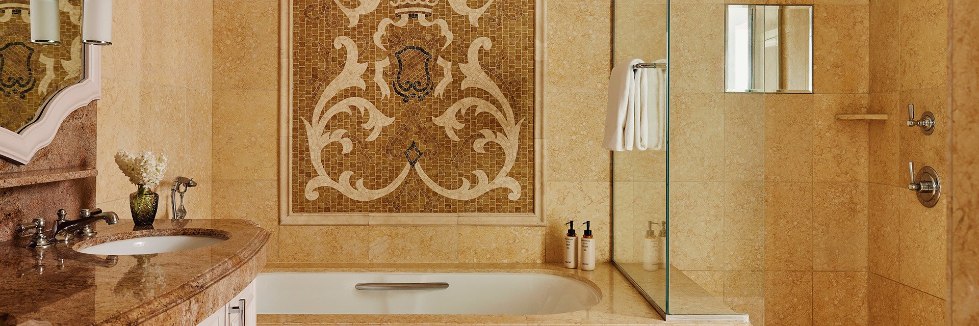 A tiled bathroom, a bathtub with a tiled wall mosaic, a hanging white towel, and a brown marble sink with white drawers.