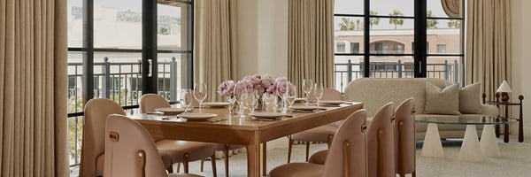 Dining table with pink potted flowers in the living area of a hotel room. Large windows let in light to an ivory couch.