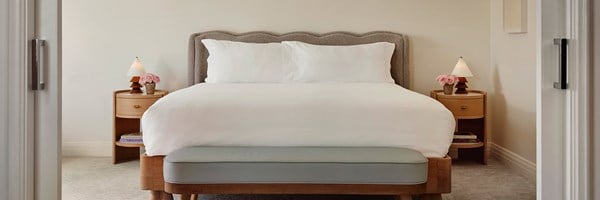 A king sized bed with white sheets. There are two wooden night stands with lamps and potted pink flowers.