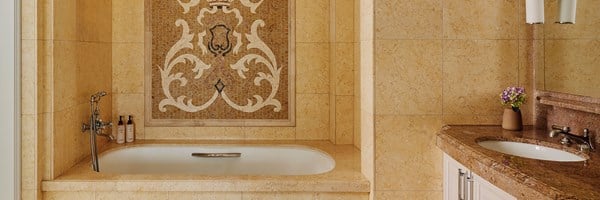 The bathroom of a hotel. The bathtub has a wall with mosaic tiles. There's a brown marble sink with purple potted flowers.