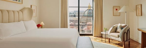 A king sized bed with white sheets. Glass doors lead out to a terrace with pink furniture. A sitting chair in the corner.