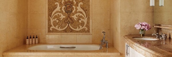 The bathroom of a hotel. The bathtub has a wall with mosaic tiles. There's a brown marble sink with pink potted flowers.