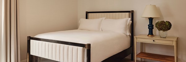A king sized bed with white sheets. There's a wooden nightstand with a lamp and potted green flowers.