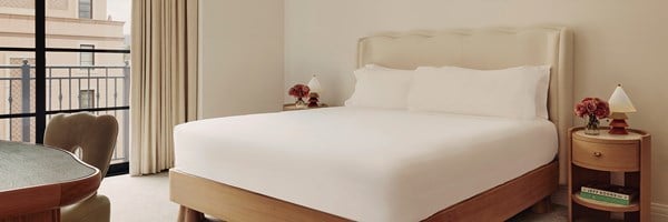 A king sized bed with white sheets. Two nightstands with lamps and violet potted roses.