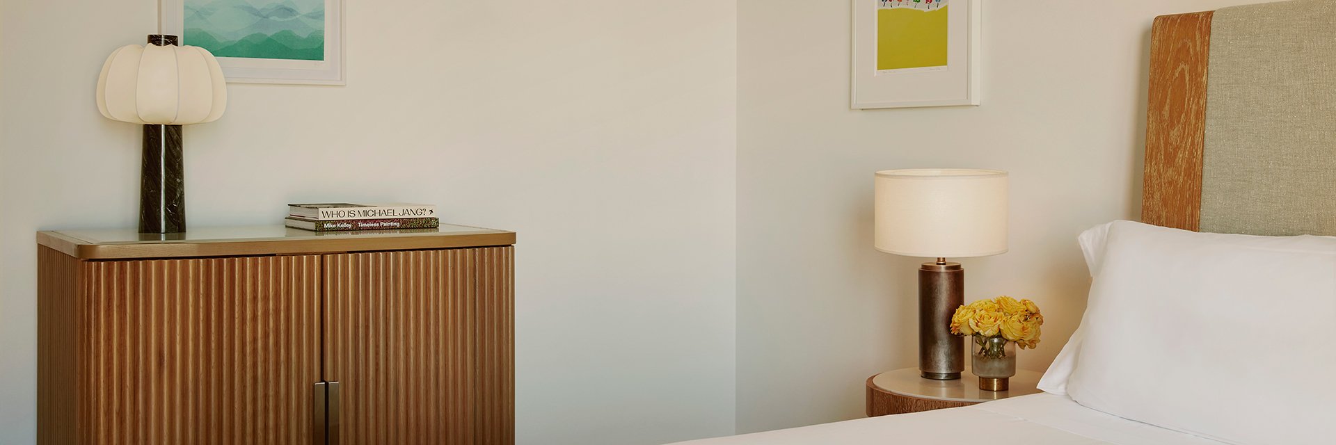 A small, wooden credenza with a lamp and two books. There are yellow roses on a nightstand beside a bed with white sheets.