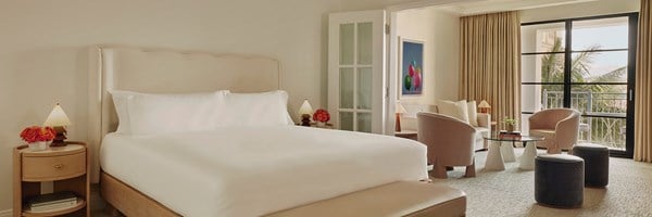 A large bed with white sheets.  A nightstand with red flowers and a small lamp. A living space with table and chairs.