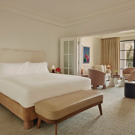 A large bed with white sheets. A nightstand with red flowers and a small lamp. A living space with table and chairs.