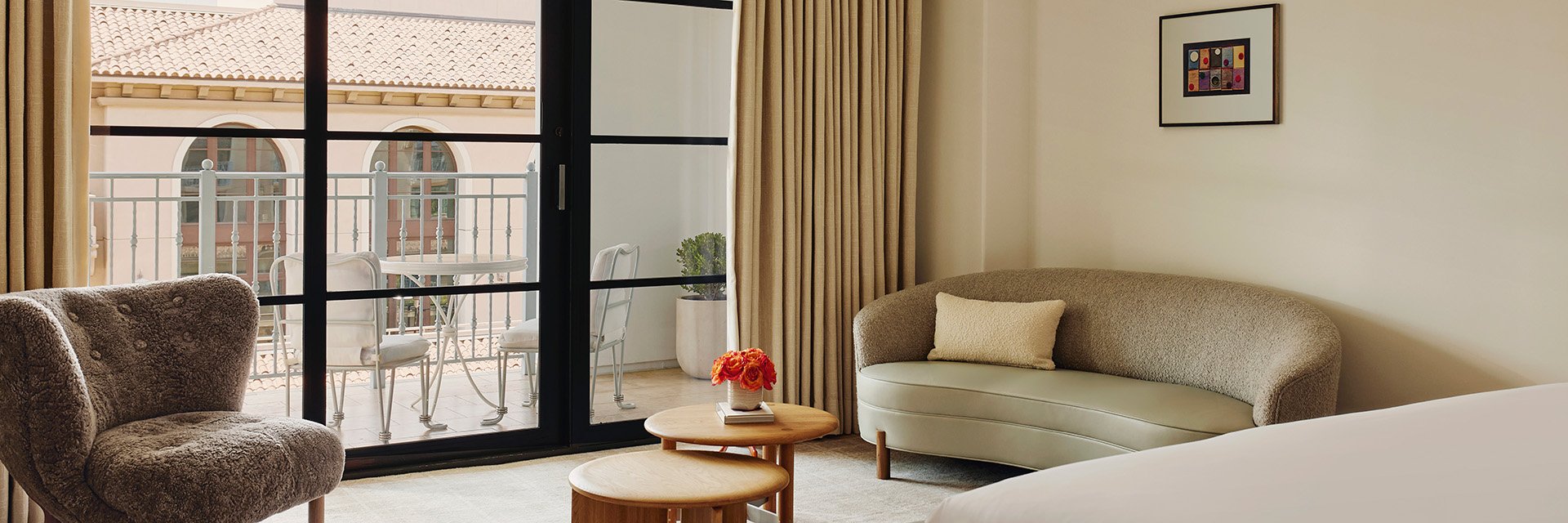A wooden coffee table with red potted flowers. There's a curved sofa and a sitting chair in front of sliding glass doors.