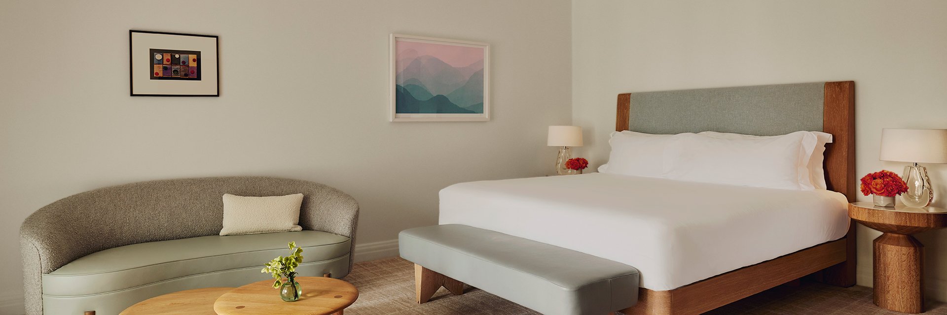 A tidy king bed with white sheets. A curved sofa. A wooden coffee table. Two nightstands with red potted roses and lamps.