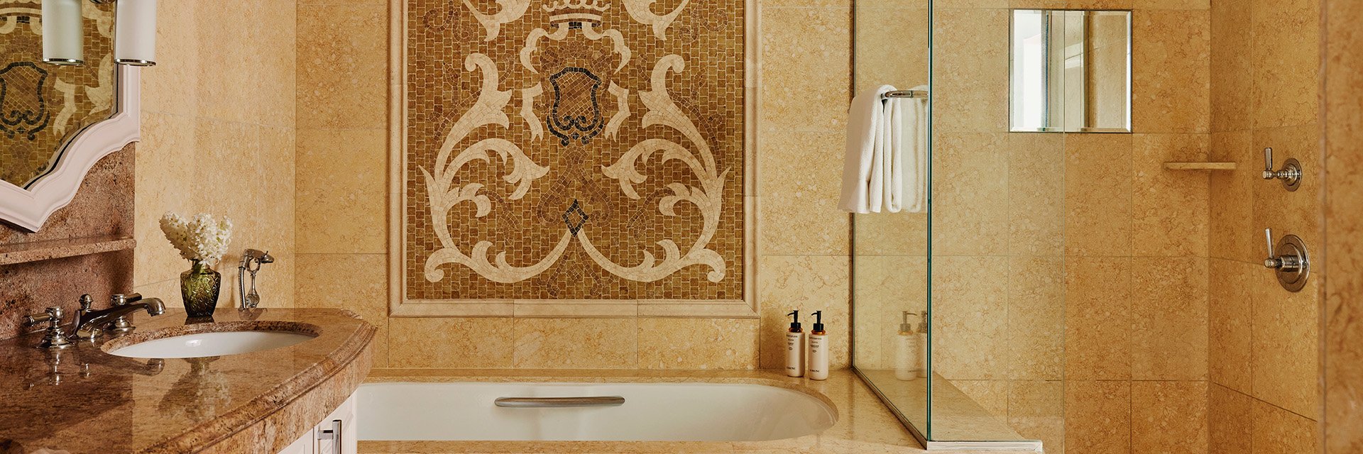 A tiled bathroom, a bathtub with a tiled wall mosaic, a hanging white towel, and a brown marble sink with white drawers.