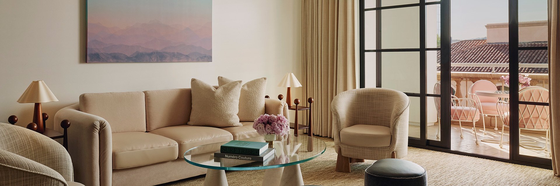 A living space with a tan couch and an ivory chair. A glass table with pink potted flowers. Open glass doors to a terrace.