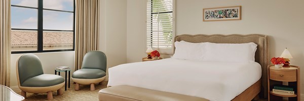 A king sized bed with white sheets. Two night stands with lamps and red potted flowers. Two mid-century blue sitting chairs.