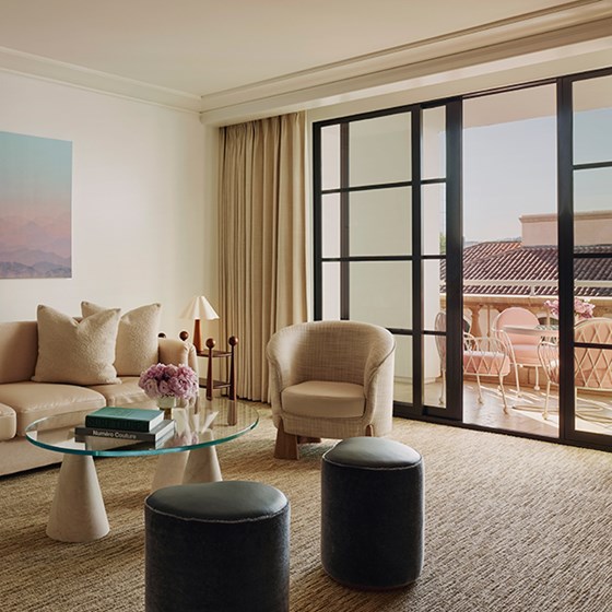 A living space with a tan couch and an ivory chair. A glass table with pink potted flowers. Open glass doors to a terrace.