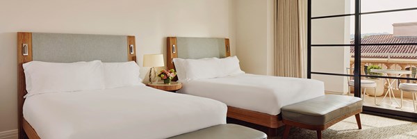 Two queen beds with white sheets. A wooden nightstand with potted flowers and a lamp. Glass doors to a terrace.