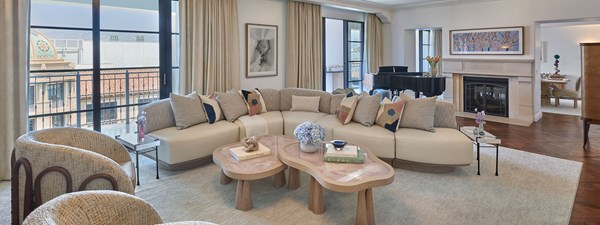 The living area of a suite. There are large windows, a curved beige couch, and two chairs. In the background is a blue piano and fireplace.