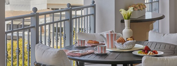 A table set on the balcony of a hotel room. There are pastries in a basket, sliced fruit on a plate, and a pink and white striped tea set.