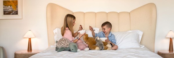 A young boy and girl sitting on a hotel bed eating from white bowls. They are surrounded by stuffed animals.