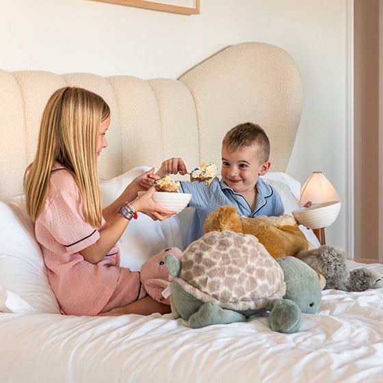 A young girl and boy sitting on a hotel bed in pajamas eating cake from bowls surrounded by stuffed animals.