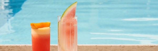 One pink watermelon cocktail and one orange cocktail lined up by pool