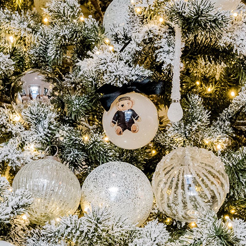 A close-up view of a frosted Christmas tree adorned with elegant ornaments, including a glass bauble featuring a teddy bear in a black bowtie.