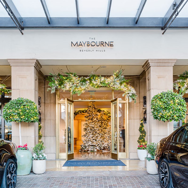 The grand entrance of The Maybourne Beverly Hills, adorned with festive greenery and sparkling decorations, featuring a majestic Christmas tree visible through the doors
