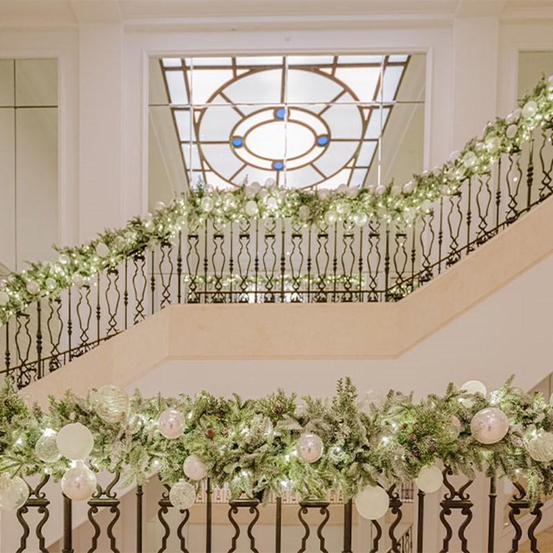 A beautifully decorated staircase wrapped in lush garlands of greenery, white ornaments, and twinkling lights under a stained glass ceiling.