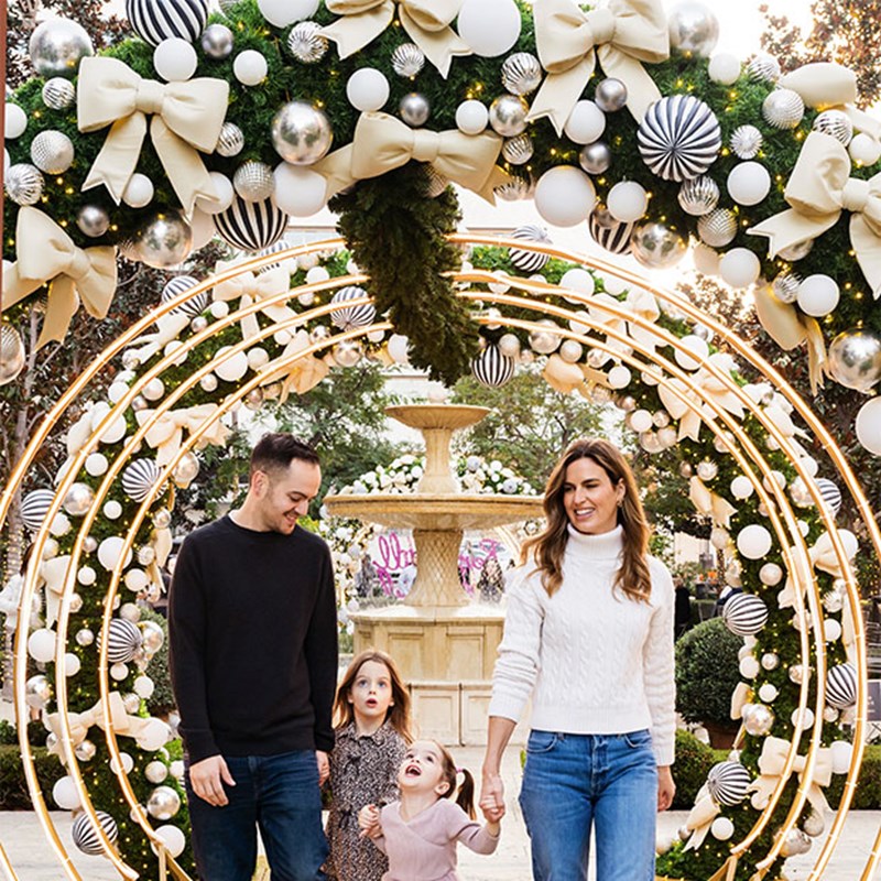 A joyful family walking hand-in-hand under an enchanting holiday arch adorned with bows, baubles, and warm lights, with a fountain in the background.