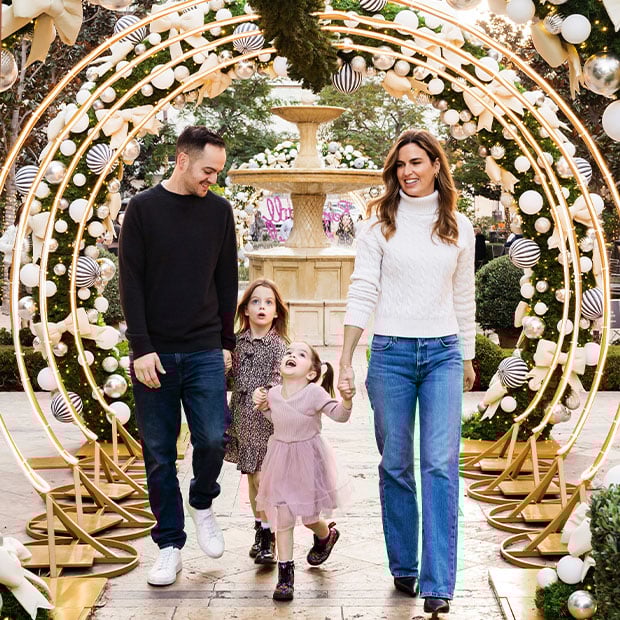 A family walks joyfully under a golden holiday arch adorned with ornaments, with a fountain and festive decorations in the background.