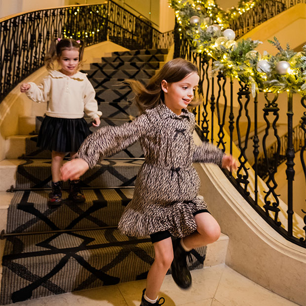 Two young sisters playfully run down a staircase adorned with elegant holiday garlands and ornaments.